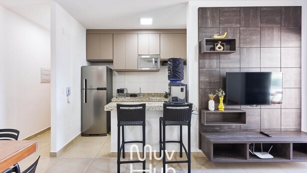 A modern kitchen with a built-in microwave, a refrigerator, and a water dispenser. There is a dining area with two chairs and a table. A flat-screen TV is mounted on a dark wall, and a shelf with decorative items is visible. - New apartment with air conditioning - REF JS45H (Ubatuba)