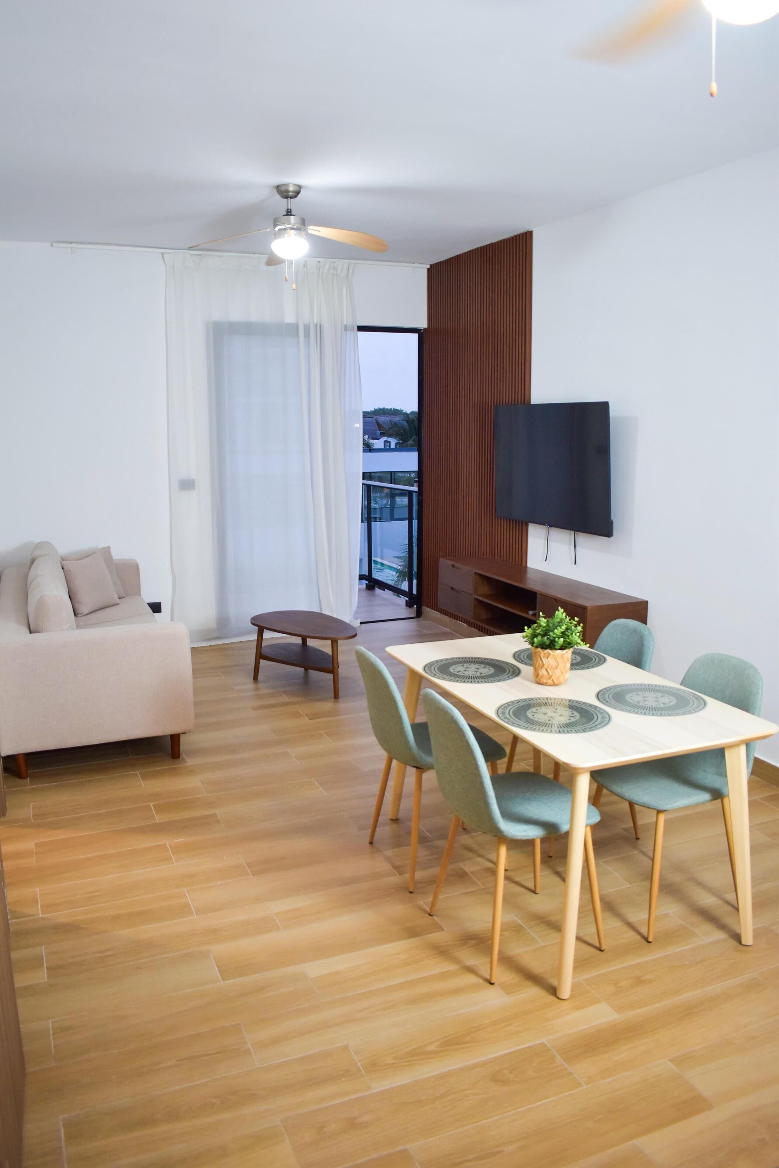 Una sala de estar moderna con un sofá, una mesa de comedor, sillas, un ventilador de techo y un televisor montado en la pared.
