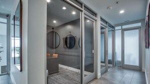 A modern hotel room with a glass door leading to a balcony, a small seating area with cushions, and a large window providing a view of the outdoors. - Cozy Comfort1bd Attached Garage+Desk+Pool+Gym (Fort Worth)