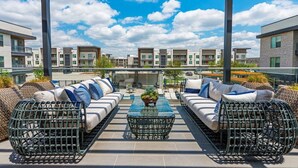 A rooftop patio with wicker furniture, a glass-top table, and a view of modern apartment buildings. - Cozy Comfort1bd Attached Garage+Desk+Pool+Gym (Fort Worth)