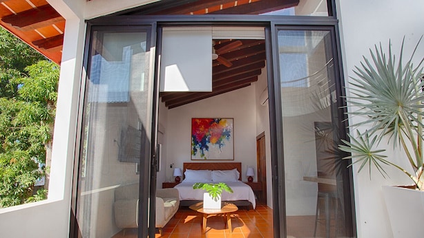 A modern hotel room with a sliding glass door, a bed with white linens, a colorful abstract painting, and a potted plant.