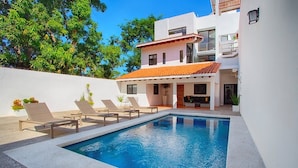 A swimming pool with lounge chairs, a covered patio, and a two-story building with a red-tiled roof.