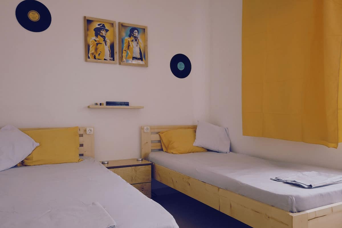 A room with two wooden beds, each with a pillow and a small bedside table. There are two framed pictures on the wall and a shelf with a book.
