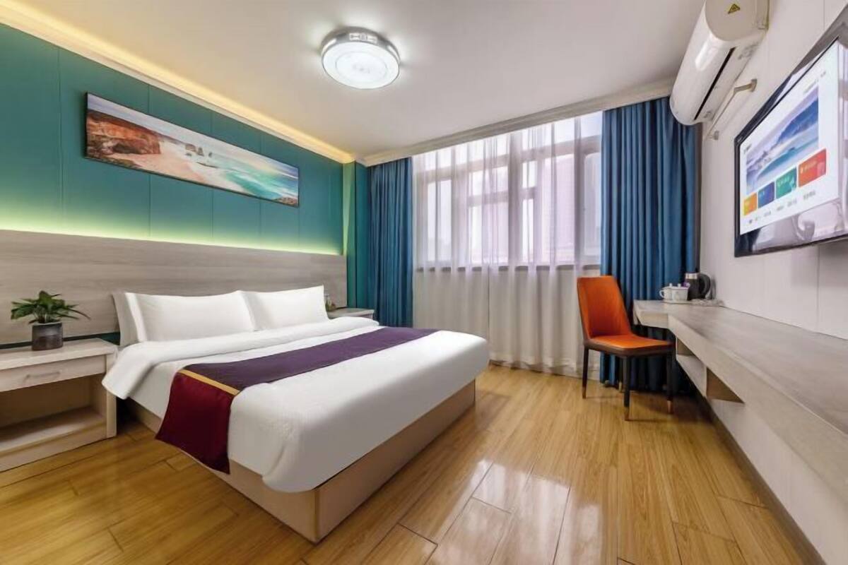 A hotel room with a bed, a desk, a chair, a window with curtains, and a wall-mounted TV.
