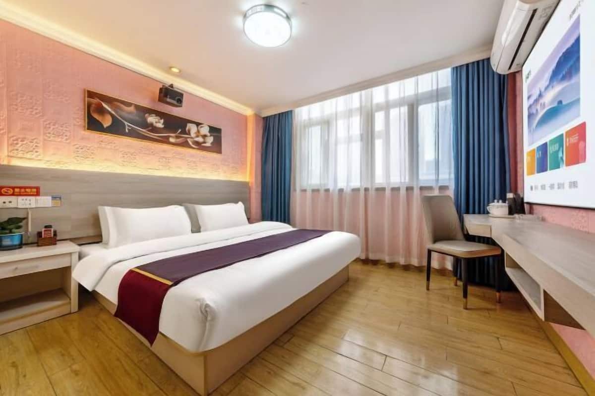 A hotel room with a bed, a desk, a chair, a window with curtains, a wall-mounted air conditioner, and a painting.