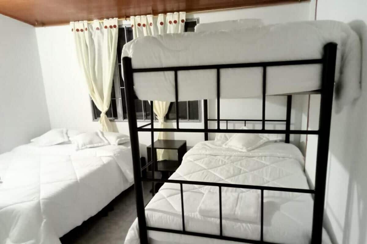 A room with two bunk beds, white bedding, a small bedside table, and a window with curtains.
