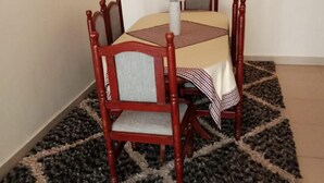A dining area with a table set for four, surrounded by wooden chairs with cushioned seats. A patterned rug covers the floor, and a framed picture hangs on the wall.
