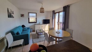 Ein Wohnzimmer mit einer Couch, einem Esstisch mit Stühlen, einem Fernseher und einem Fenster mit Vorhängen.