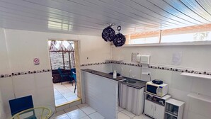 A compact kitchen with white tiles, a glass table, blue chairs, a microwave, and a small cabinet. - Pousada Golfinho (Fernando de Noronha)