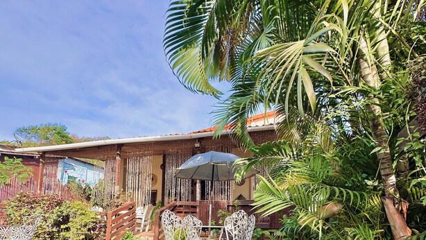 A wooden deck with outdoor seating, surrounded by lush greenery and a building with a red roof. - Pousada Golfinho (Fernando de Noronha)