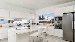 A modern kitchen with white cabinetry, a central island with bar stools, a stainless steel refrigerator, and a view of a white building with palm trees. - Villa Bolboreta by Villa Plus (Teguise)