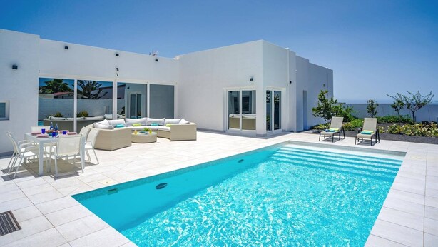 A modern outdoor pool area with a clear blue pool, white outdoor furniture, and a view of the sea. - Villa Bolboreta by Villa Plus (Teguise)