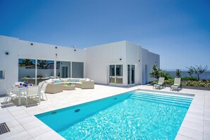 Een modern buitenzwembad met een helderblauw zwembad, witte tuinmeubels en uitzicht op de zee.