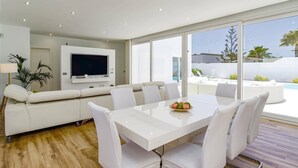 A modern living room with a white sofa, a TV mounted on the wall, and a dining area with a white table and chairs. - Villa Bolboreta by Villa Plus (Teguise)