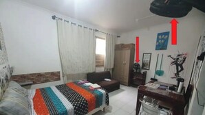 Un dormitorio con cama, escritorio y armario. Hay cortinas en la ventana y un ventilador en la pared.