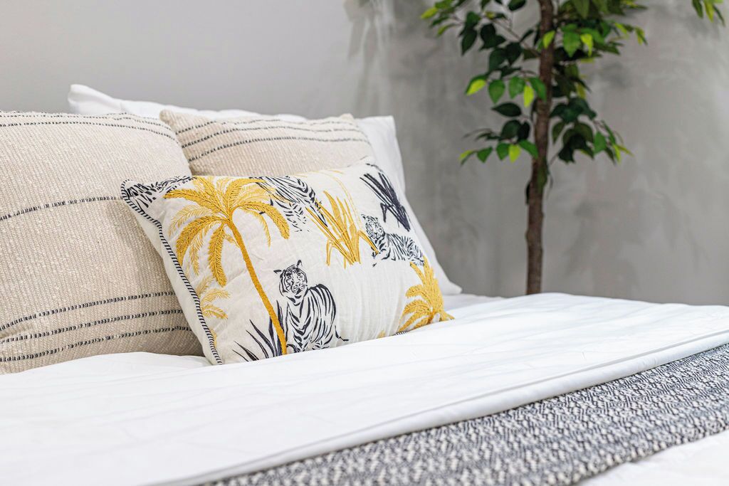 Cama com roupa de cama branca e duas almofadas, uma com padrão tropical e outra listrada.