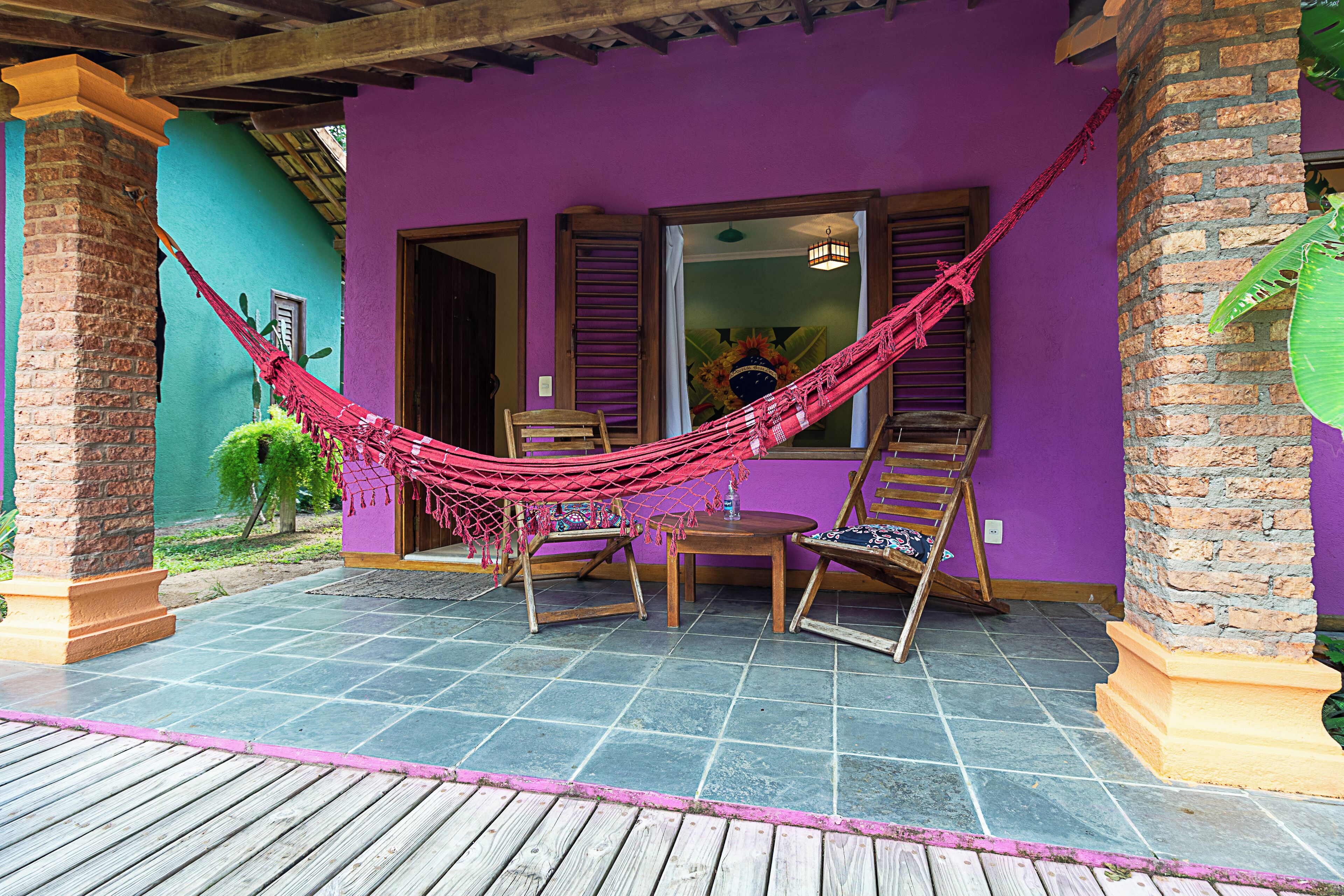 A hammock set up between two wooden chairs on a patio with a purple wall and a tiled floor.