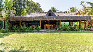 A thatched-roof structure with an open entrance, surrounded by lush greenery and palm trees. - Villa Maravilha (Porto Seguro)