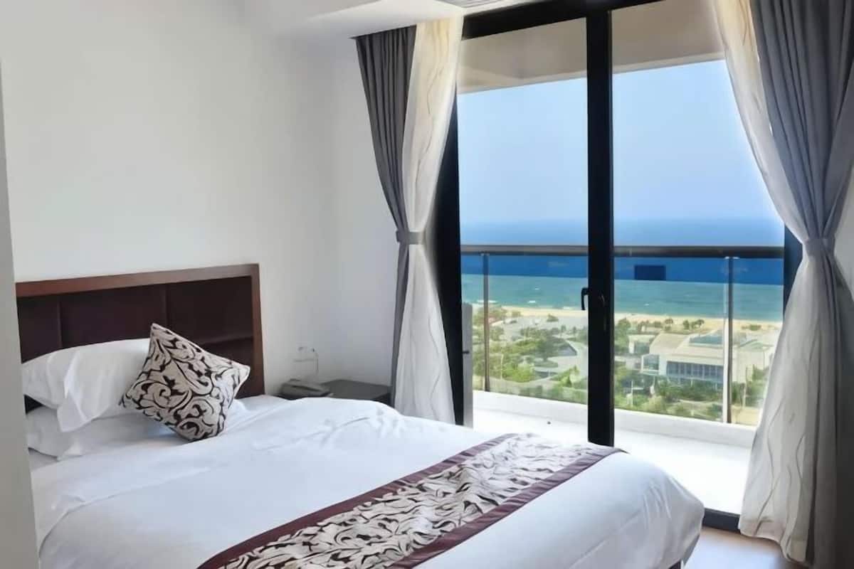 A hotel room with a bed, a nightstand, a window with curtains, and a view of the ocean.