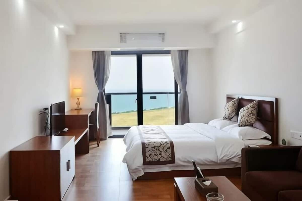 A hotel room with a bed, a desk, a TV, and a view of the sea.