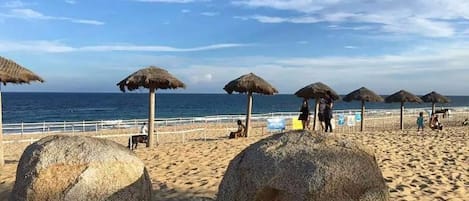 A beach with thatched umbrellas and large rocks.