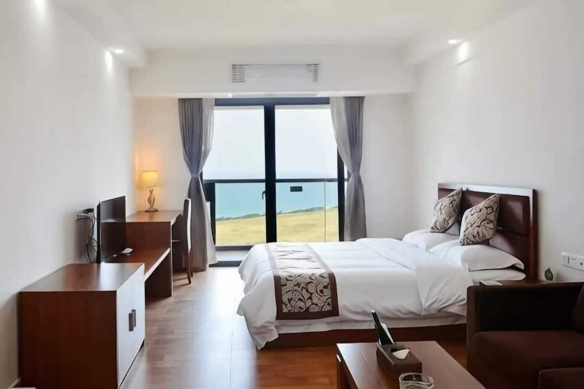 A hotel room with a bed, a desk, a television, and a view of the beach.