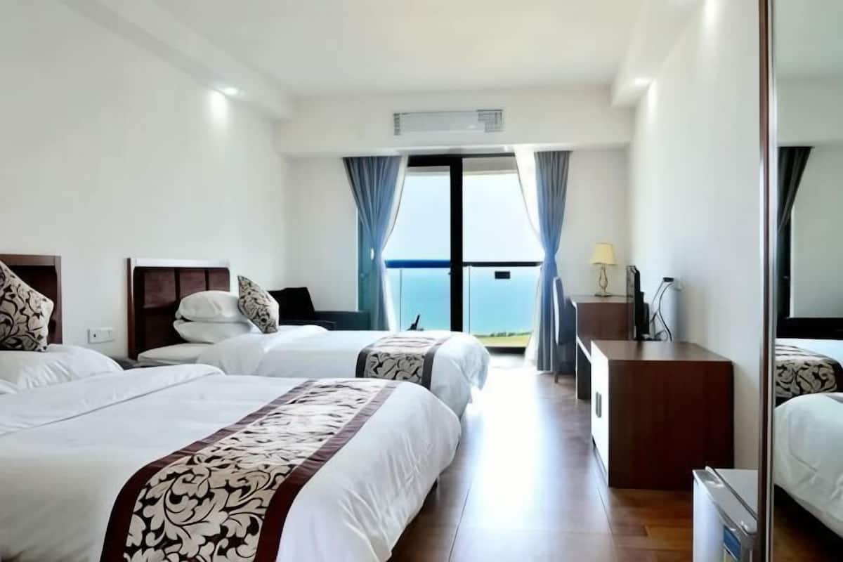 A hotel room with two beds, a desk, a chair, a lamp, and a view of the sea.