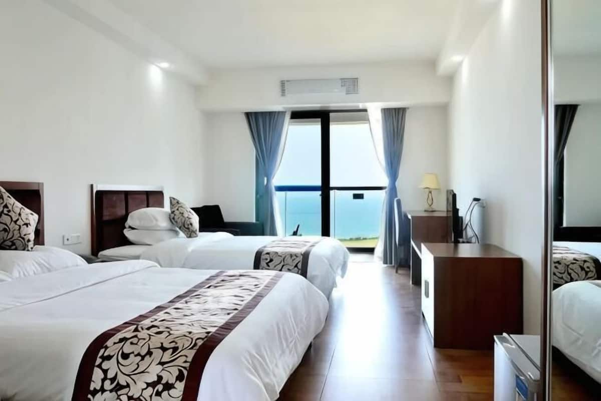 A hotel room with two beds, a desk, a chair, a lamp, and a view of the sea.