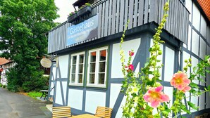 A traditional building with a sign indicating it's a guesthouse since 2023, outdoor seating, and a garden with flowers.