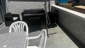 An outdoor terrace with a stone wall, a table with a patterned tablecloth, two black wicker chairs, a white chair, and a green umbrella providing shade.