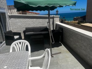 An outdoor terrace with a stone wall, a table with a patterned tablecloth, two black wicker chairs, a white chair, and a green umbrella providing shade.