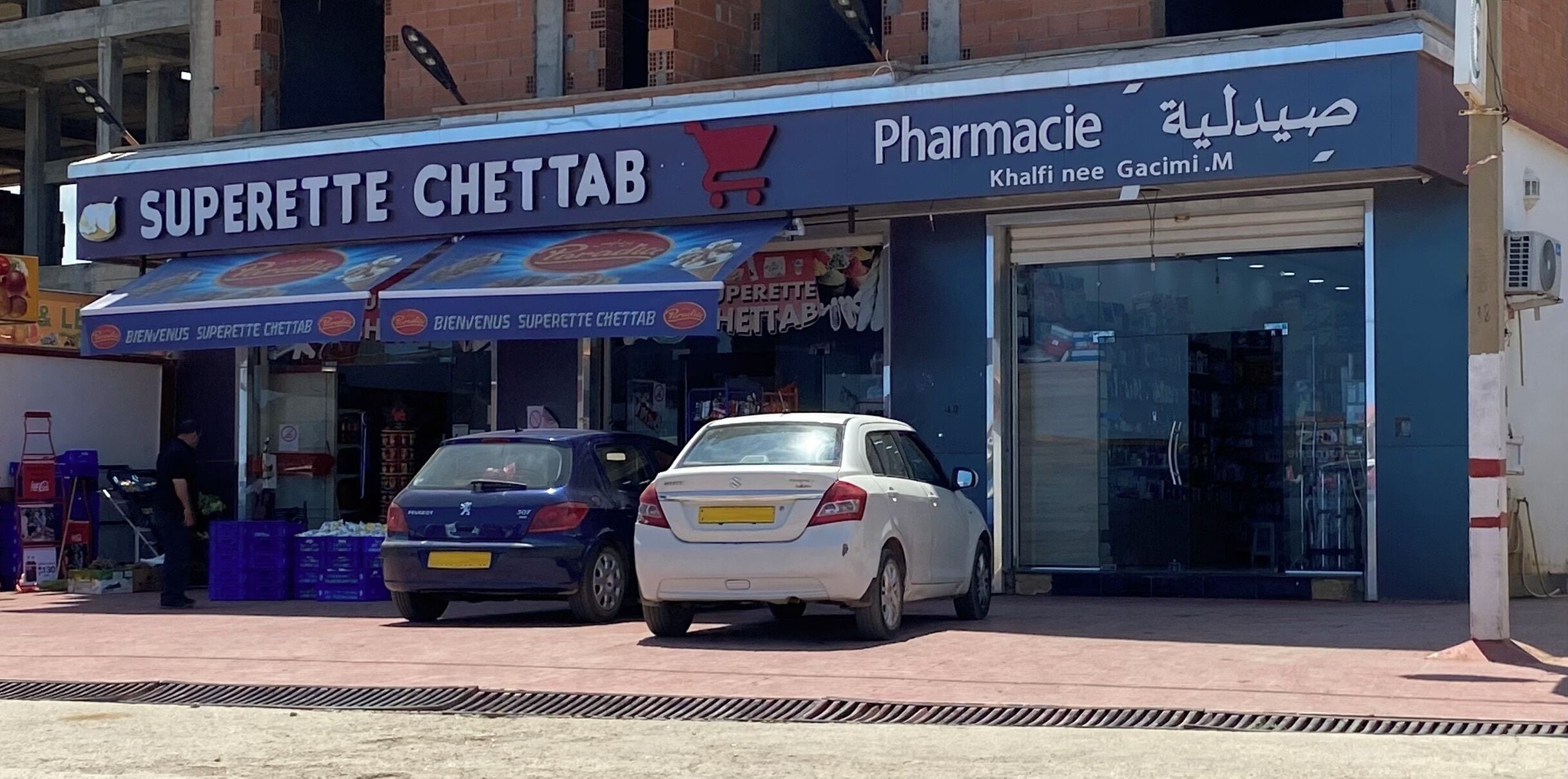 Una farmacia llamada 'Superette ChetTab' con un letrero y dos coches estacionados frente a ella.