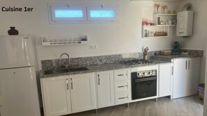 A kitchen with white cabinets, a built-in dishwasher, a gas stove, and a refrigerator. There are two windows above the sink and a shelf with jars and a kettle. - Maison Individuelle - Ait Chafaa Azeffoun (Aït Chafâa)