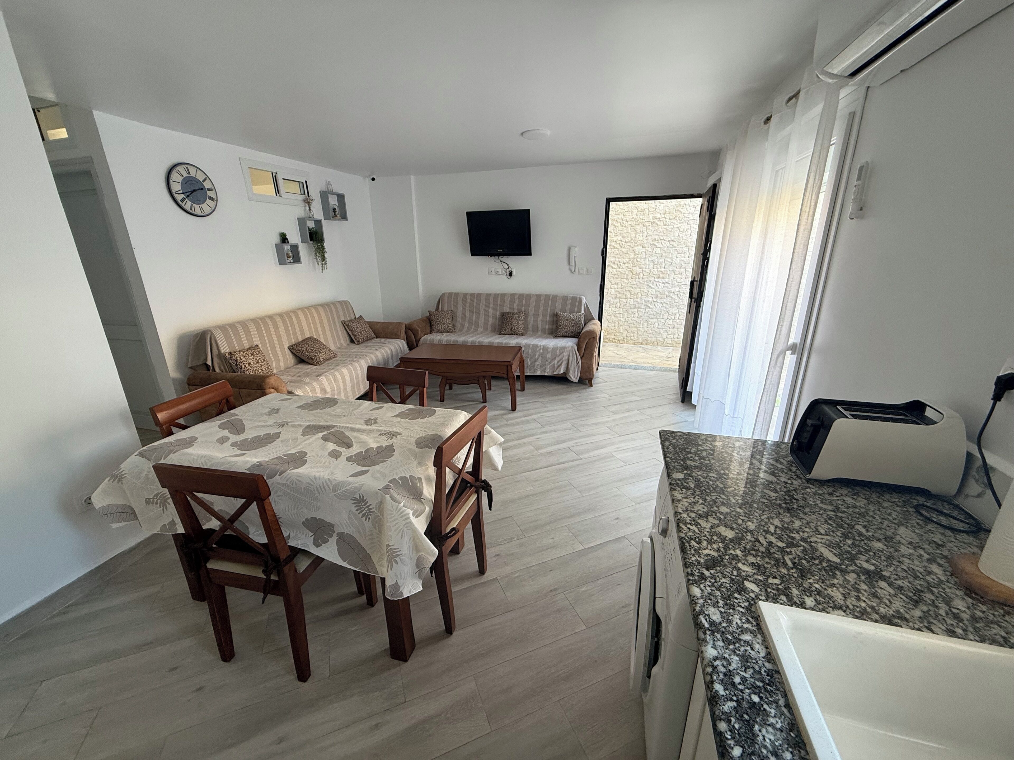 Un coin repas avec une table et des chaises, un ensemble de canapés, un téléviseur à écran plat fixé au mur et un coin cuisine équipé d’un grille-pain.