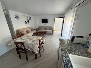 A dining area with a table and chairs, a sofa set, a flat-screen TV mounted on the wall, and a kitchen area with a toaster. - Maison Individuelle - Ait Chafaa Azeffoun (Aït Chafâa)