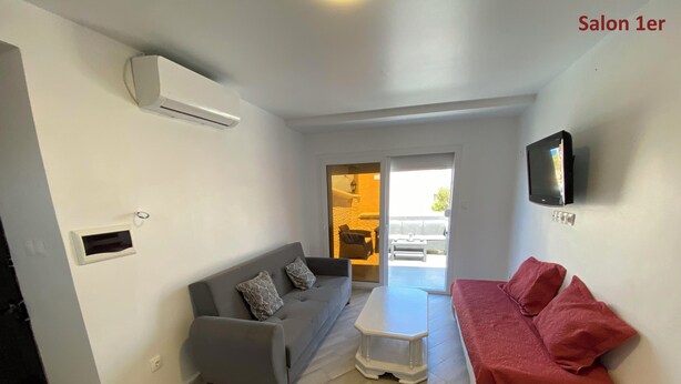 A modern living room with a grey sofa, a red sofa, a small white table, and a TV mounted on the wall. - Maison Individuelle - Ait Chafaa Azeffoun (Aït Chafâa)