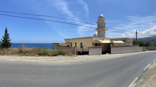 A mosque with a dome and minaret, located beside a road with a view of the sea.
