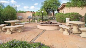 Un patio con mesas de piedra y un hogar, rodeado de follaje y un edificio al fondo.
