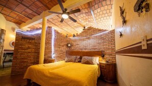 A room with a brick wall, a bed with a yellow bedspread, a ceiling fan, and a nightstand with a lamp. - Casa Happy Sol - Delfin (San Francisco)