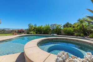 Une piscine aux eaux cristallines, dotée d’un panier de basketball, entourée d’une végétation luxuriante et délimitée par une clôture.