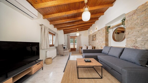 A spacious living room with a large flat-screen TV, a grey sofa, a wooden coffee table, and a stone wall. - Elida Inn (Markopoulo Mesogaias)