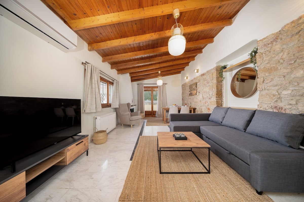 A spacious living room with a large flat-screen TV, a grey sofa, a wooden coffee table, and a stone wall.