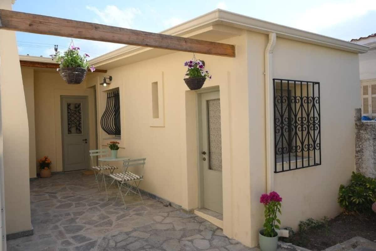 A small, cozy outdoor patio with a stone floor, a table with chairs, hanging flower baskets, and a metal gate.