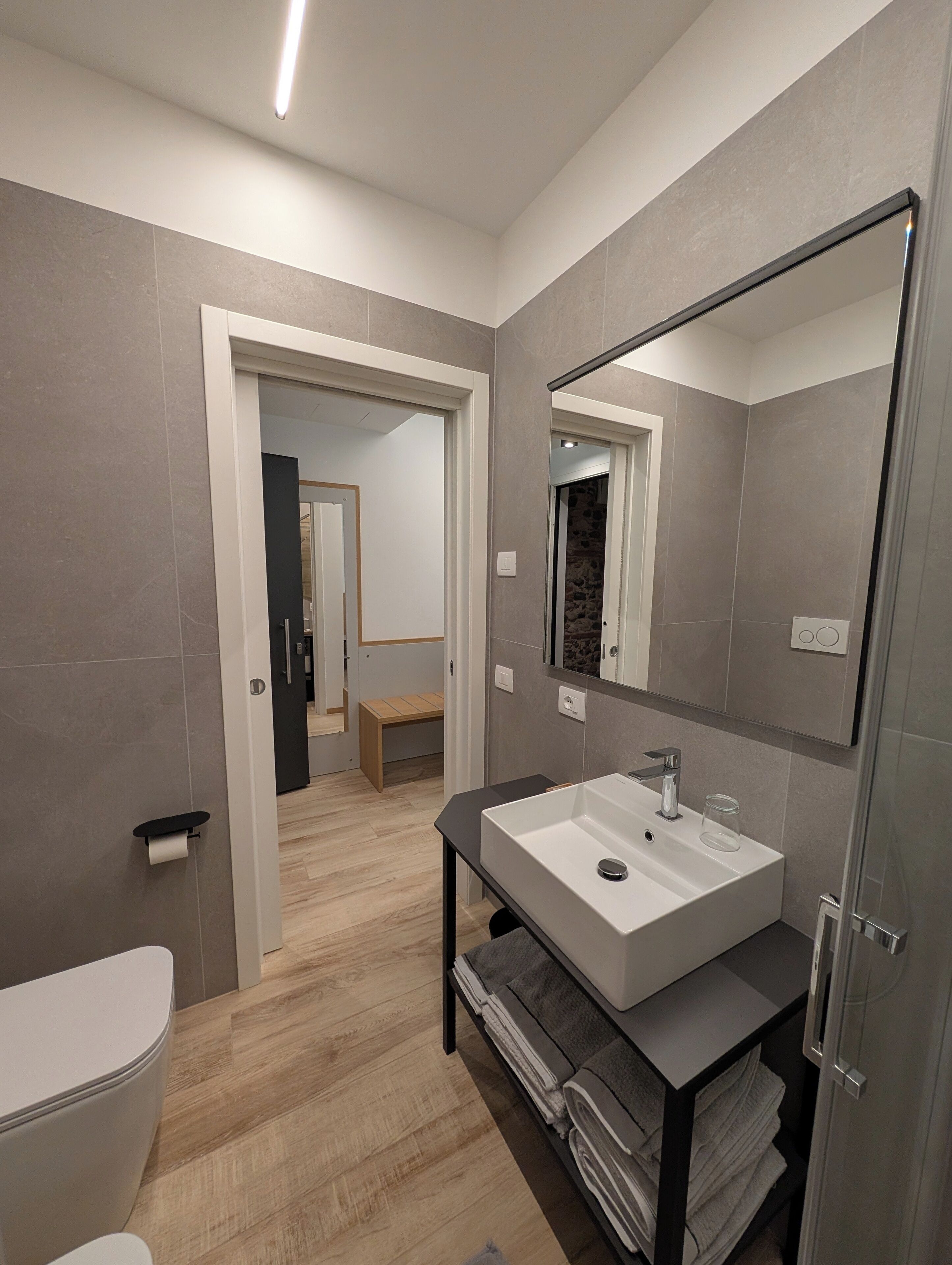 A modern bathroom with a white sink, a large mirror, and a wooden bench in the background.