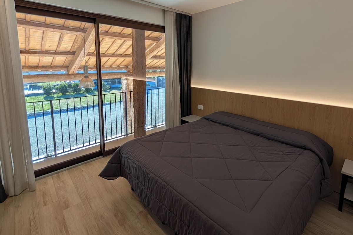 A modern bedroom with a large bed, wooden flooring, a balcony with a view of a park, and a wooden ceiling structure.