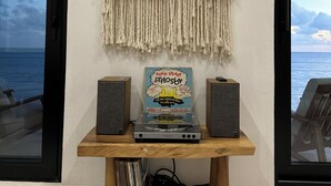Een wanddecoratie van macramé, een platenspeler en twee speakers op een houten tafel, met uitzicht op de oceaan tegen een witte muur.