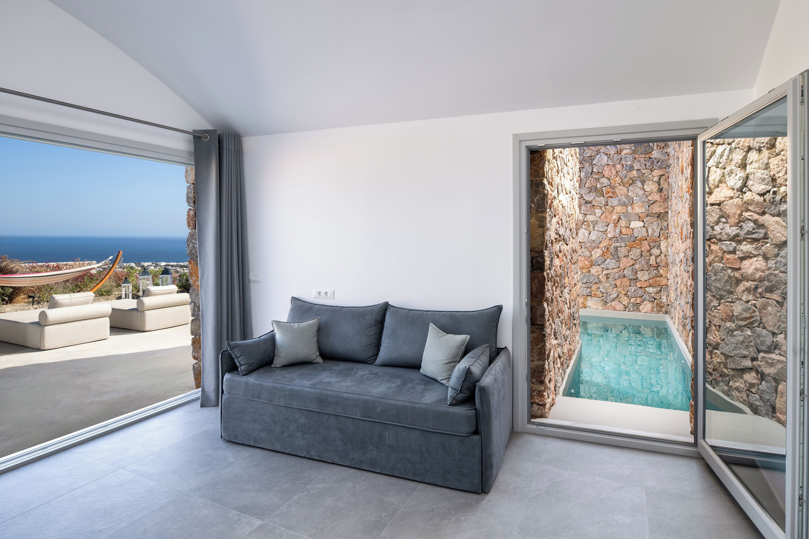 Una sala de estar moderna con un sofá gris, una pared de piedra y un ventanal que da a un balcón con vistas al mar.