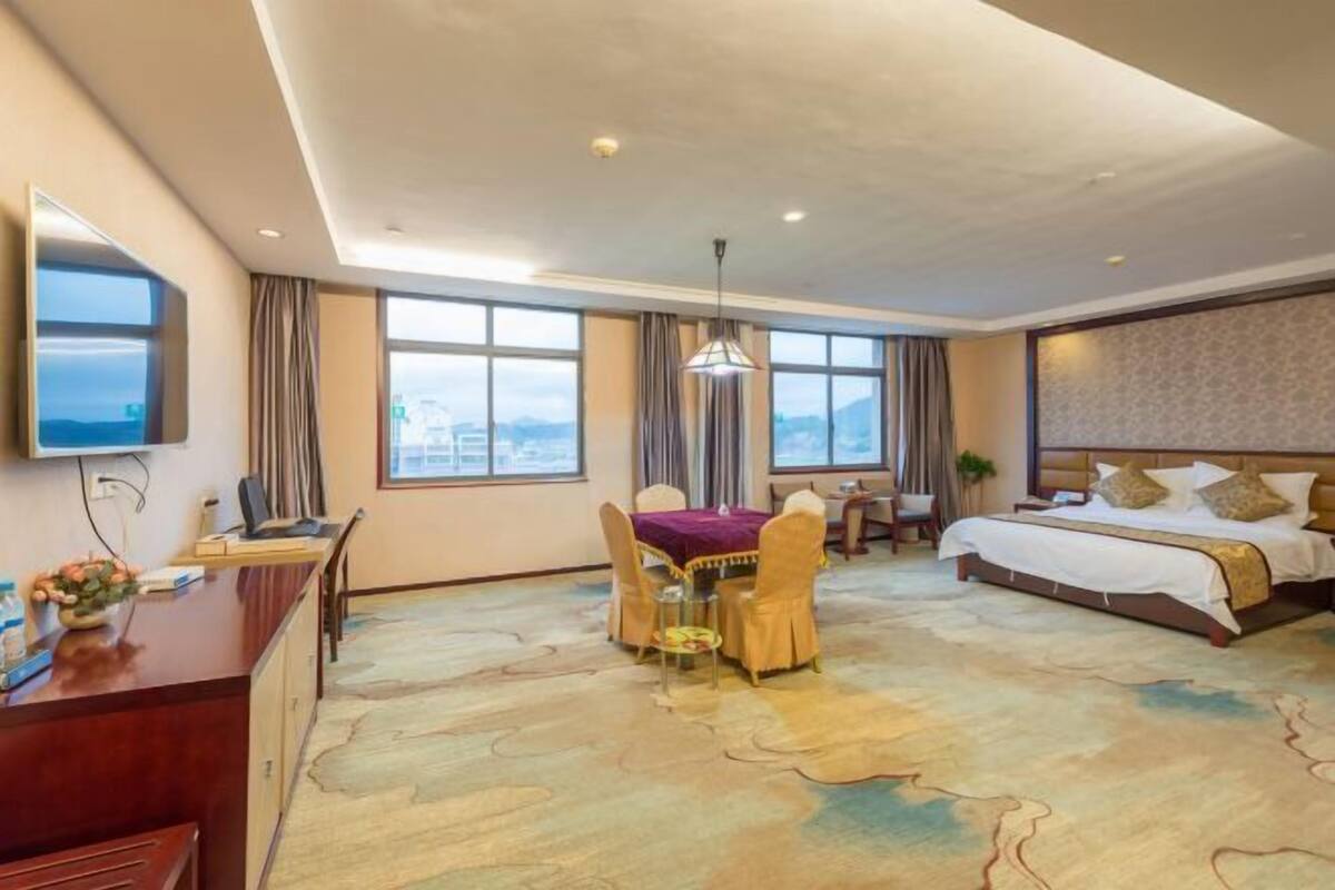 A hotel room with a bed, desk, chair, dining table, and large windows.