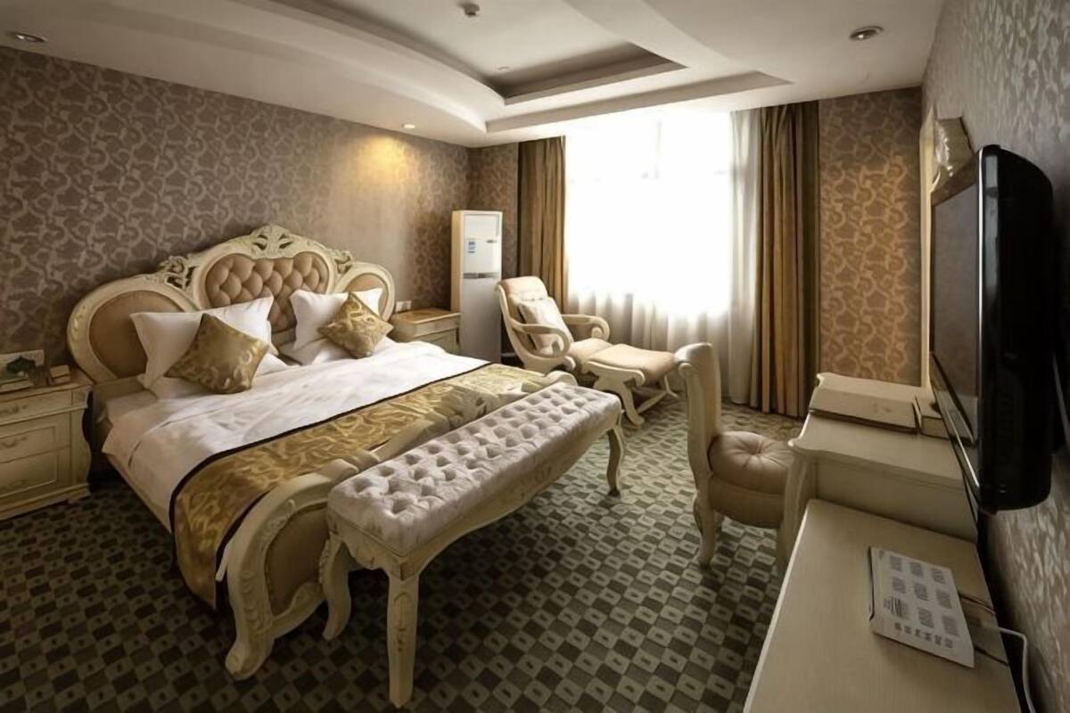 A hotel room with a large bed, a desk with a television, a chair, and a bench.