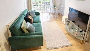 A living room with a green sofa, a flat-screen TV, and a dining area with white chairs and a glass table. - Modern 2BR Garden Flat King’s Cross - London (Greater London)